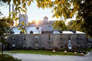 Sunset view of a stone-built distillery with a metal roof, surrounded by trees with whiskey barrels along the path.